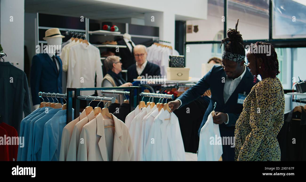 afroamerikanische Arbeiterin zeigt schwangere Frauen weiße Hemden und hilft ihr dabei, die richtige Größe für den Rabatt zu wählen. Ladenassistent empfiehlt Kleidung von Designermarken. Kamera B.. Stockfoto