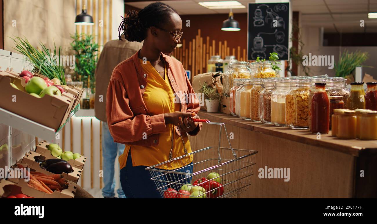 Stammgast auf der Suche nach umweltfreundlichen Alternativen für Lebensmittel, die sich hausgemachte ethisch produzierte Massenartikel in einem Supermarkt vor Ort anschauen. afroamerikanische Veganerin geht einkaufen. Kamera 2. Stockfoto