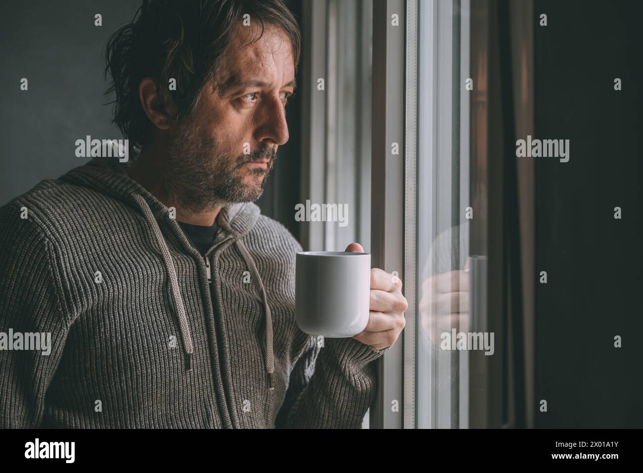 Nachdenklicher männlicher Erwachsener trinkt Kaffee am Morgen am Fenster des Appartements, während er hinausschaut und nachdenkt. Coffeindosis zum Tagesbeginn. Wählen Sie Aus Stockfoto