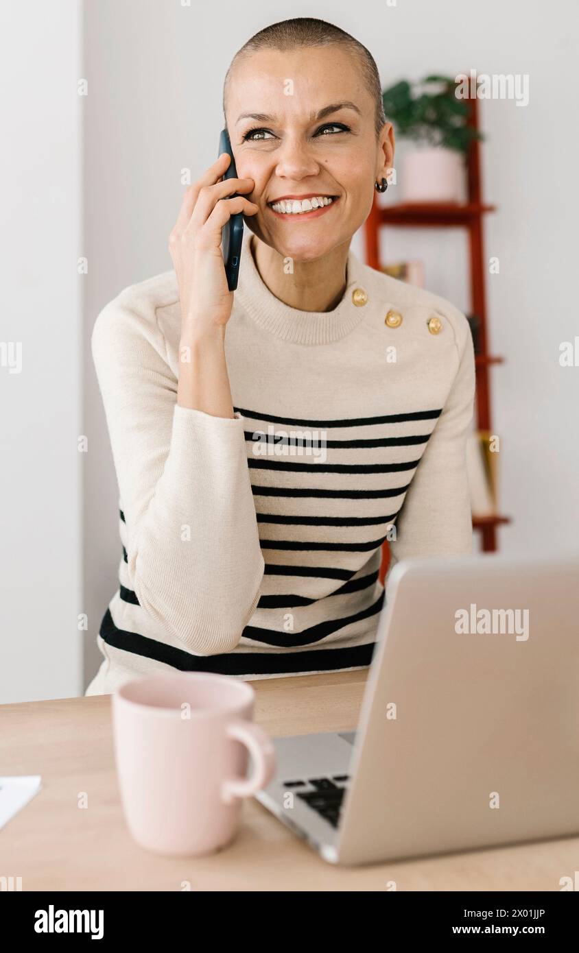 Glückliche mittelErwachsene Frau, die auf dem Handy spricht und am Arbeitsplatz sitzt Stockfoto