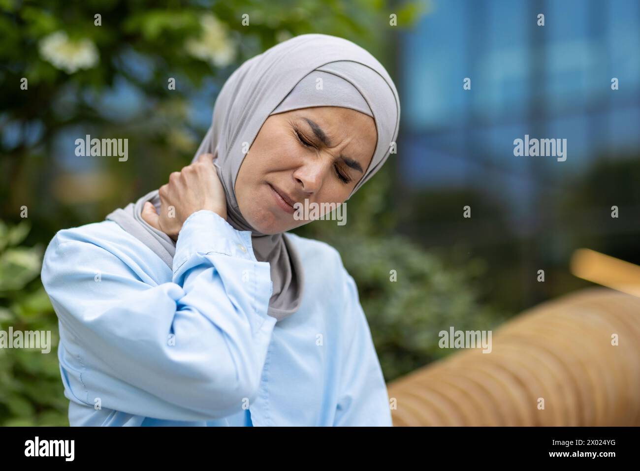 Porträt einer ungesunden arabischen Dame, die den Kopf mit geschlossenen Augen beim Reiben des Nackens auf verschwommenem Hintergrund beugt. Kranke Frau mit Entzündungen in Muskeln und Schultermassagen als tägliche Therapie im Freien. Stockfoto