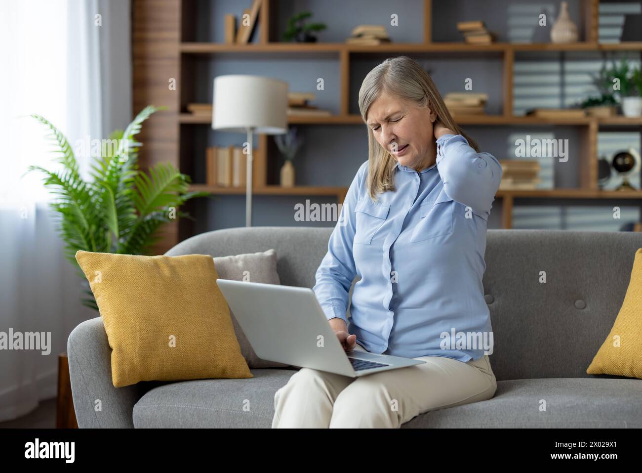 Müde reife Dame in blauem Hemd, die sich den Nacken reibt, während sie auf der Couch sitzt und einen Laptop auf den Beinen hat. Überlastete Frau, die den Hinterkopf mit geschlossenen Wows berührt, um die Belastung der Wirbelsäulenmuskulatur zu entlasten. Stockfoto