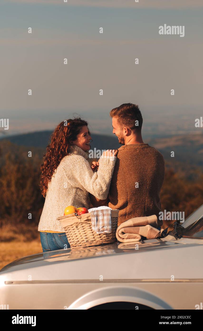 Rückansicht eines Paares, das bei Sonnenuntergang Picknick genießt. Sie sitzen auf einem altmodischen Auto und schauen auf die Distanz. Stockfoto