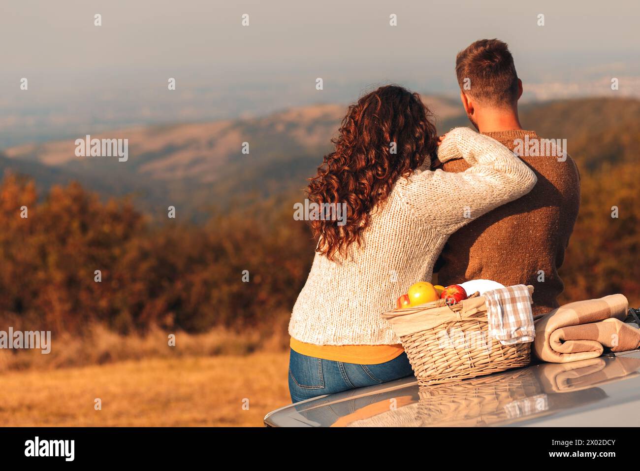 Rückansicht eines Paares, das bei Sonnenuntergang Picknick genießt. Sie sitzen auf einem altmodischen Auto und schauen auf die Distanz. Stockfoto