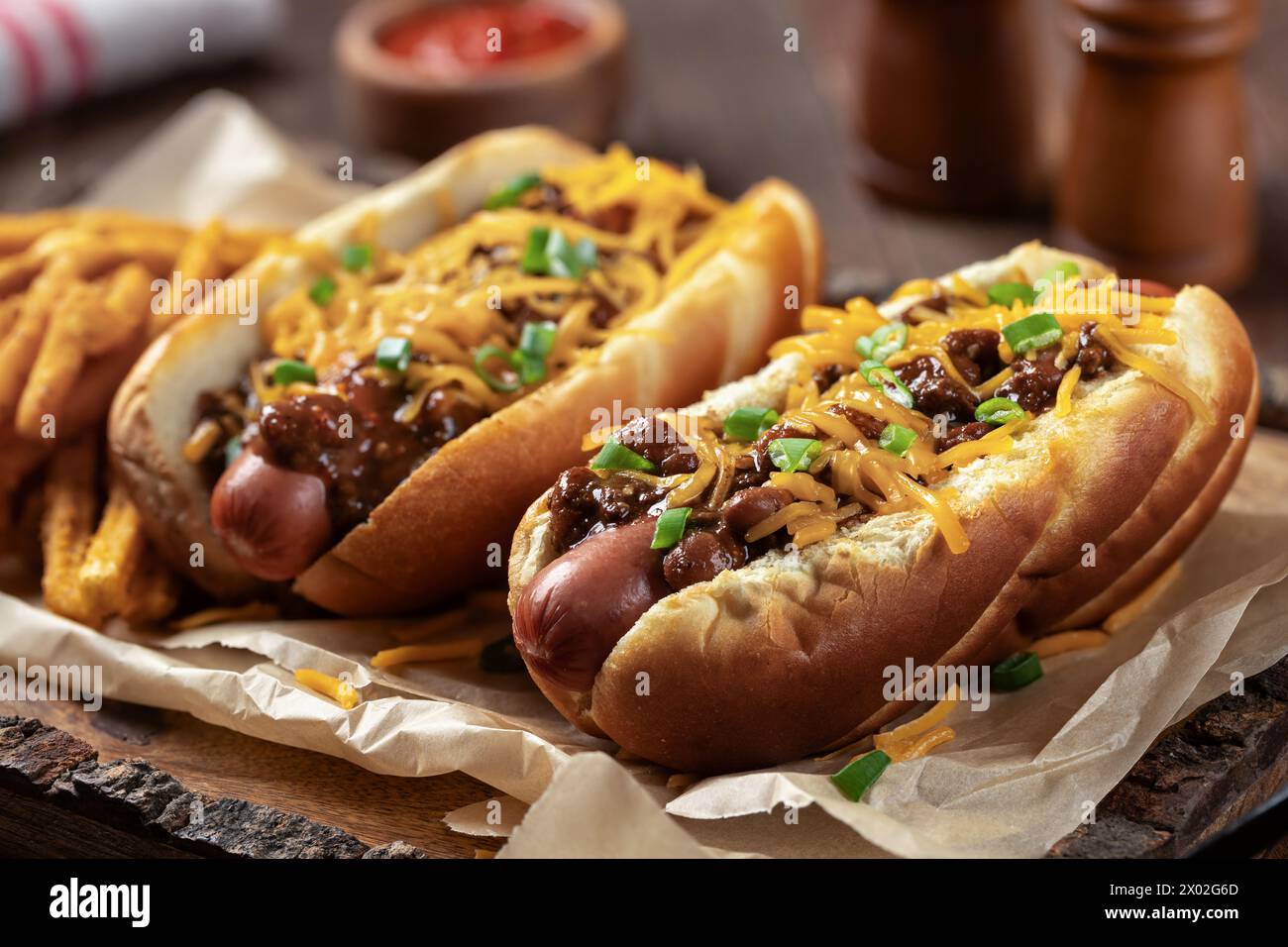 Zwei Chili Hot Dogs mit geriebenem Cheddar, gehackten grünen Zwiebeln und gewürzten Pommes frites auf einem Holztablett Stockfoto