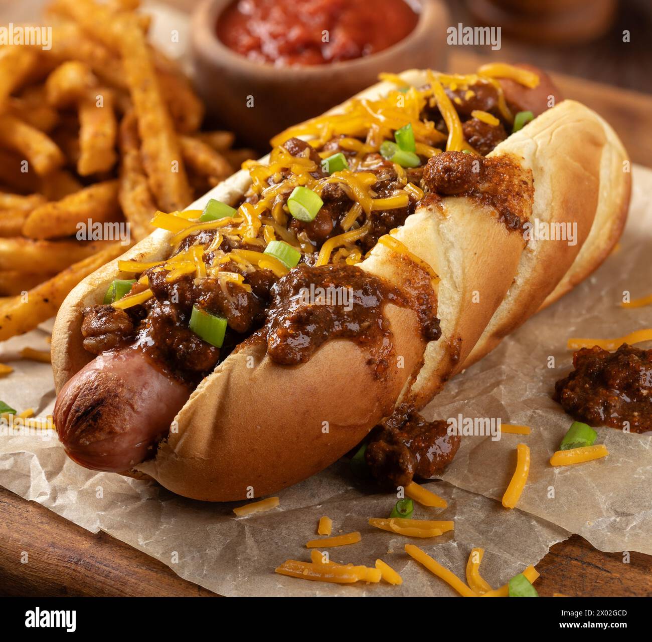 Chili Hot Dog mit geriebenem Cheddar und gewürzten Pommes frites auf einem Holzbrett Stockfoto