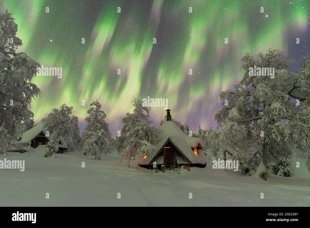 Winterblick auf eine typische Holzhütte, beleuchtet durch das Feuer im gefrorenen Wald während eines Nordlichtersturms (Aurora Borealis), Finnisch-Lappland, Finnland Stockfoto