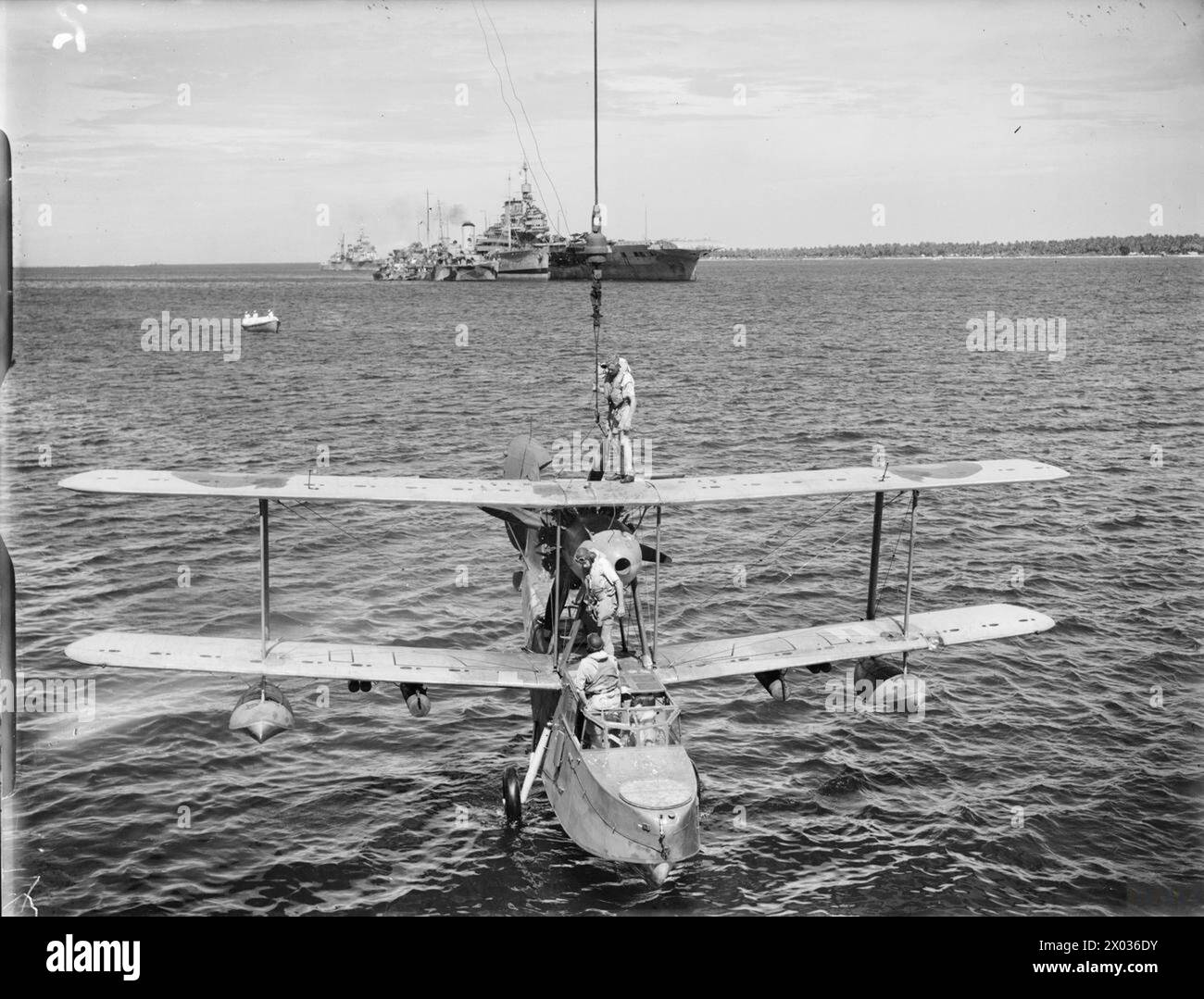 DIE ROYAL NAVY WÄHREND DES ZWEITEN WELTKRIEGS – Ein Amphibienflugzeug der Supermarine Walrus wird nach einer Rückfahrt im Indischen Ozean an Bord der HMS WARSPITE gehoben. Die Besatzungsmitglieder stehen sowohl auf dem oberen als auch auf dem unteren Flügel und halten das Flugzeug während des Aufzugs. Im Hintergrund ist ein BERÜHMTER Flugzeugträger zu sehen. Neben ihr befindet sich ein weiteres Schiff, möglicherweise ein Krankenhausschiff, und die HMS LUDLOW. Ein Kreuzer ist hinter diesen Schiffen verankert und ein Motorschneider macht sich auf den Weg von dieser Gruppe von Schiffen in Richtung der Kamera Royal Navy, WARSPITE (HMS) Stockfoto