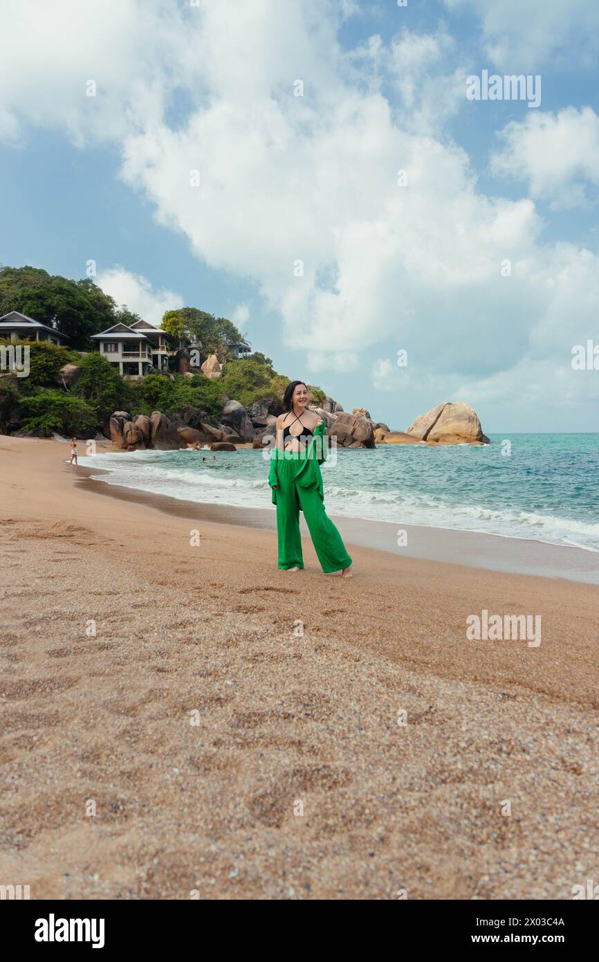 Stilvolle Frau macht einen gemütlichen Spaziergang entlang eines ruhigen Strandes, während das Meer sanft am Ufer liegt Stockfoto