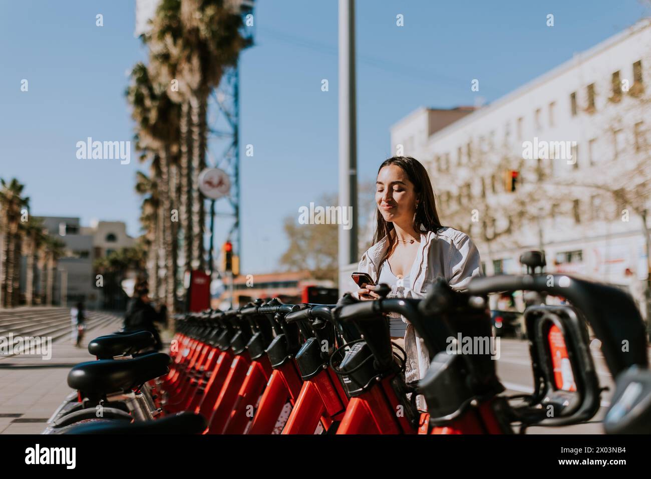 Die junge Frau steht bei einer Reihe roter Fahrräder im sonnigen Barcelona und überprüft ihr Handy, wahrscheinlich mit einer Bike-Sharing-App Stockfoto