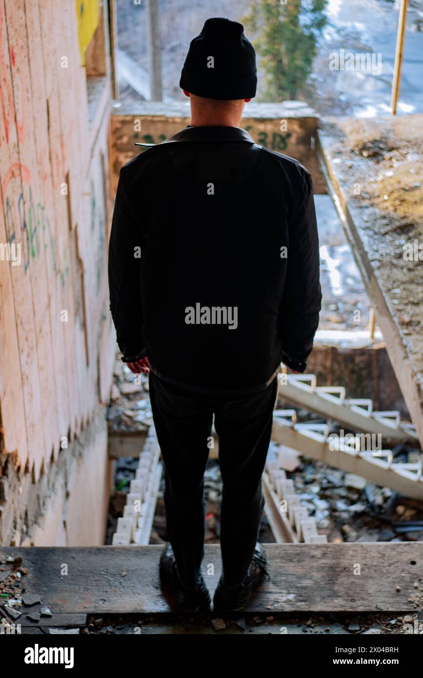 Ein junger Mann in einer schwarzen Lederjacke steht mit dem Rücken zur Kamera vor dem Hintergrund einer zerstörten Treppe in einem verlassenen Gebäude. Stockfoto