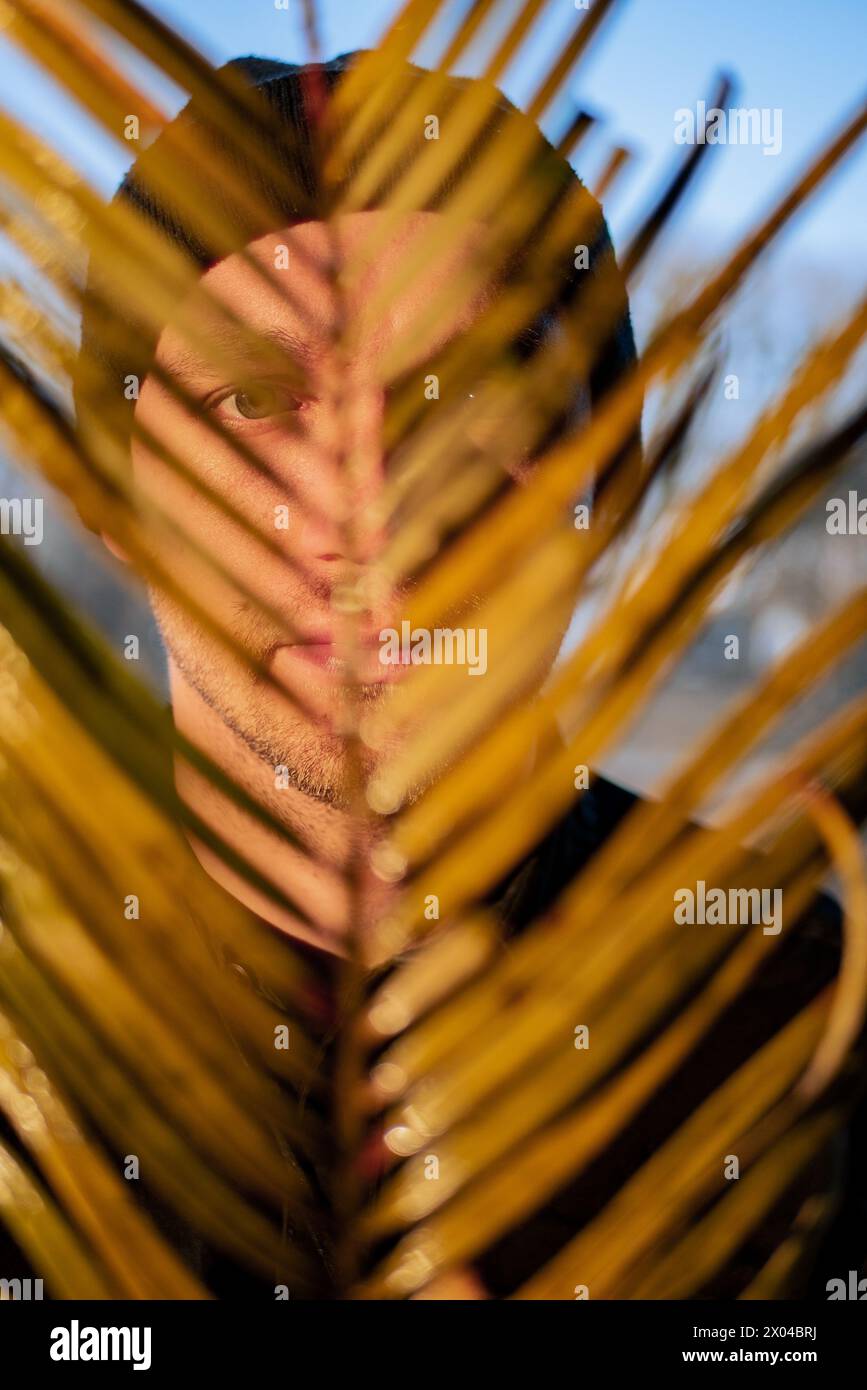 Porträt eines jungen gutaussehenden Typen mit schwarzem Hut, der sein Gesicht mit trockenen Palmblättern bedeckt. Stockfoto