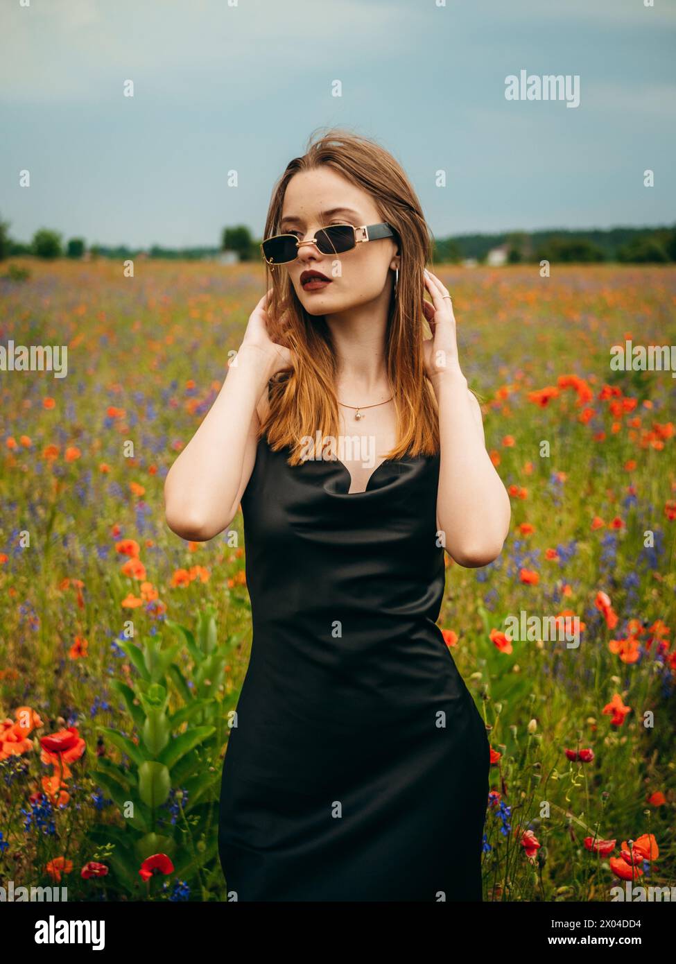 Schönes junges Mädchen in einem schwarzen Abendkleid und einer Sonnenbrille, die an einem bewölkten Sommertag vor einem Mohnfeld posiert. Porträt eines weiblichen Modells im Freien. Stockfoto