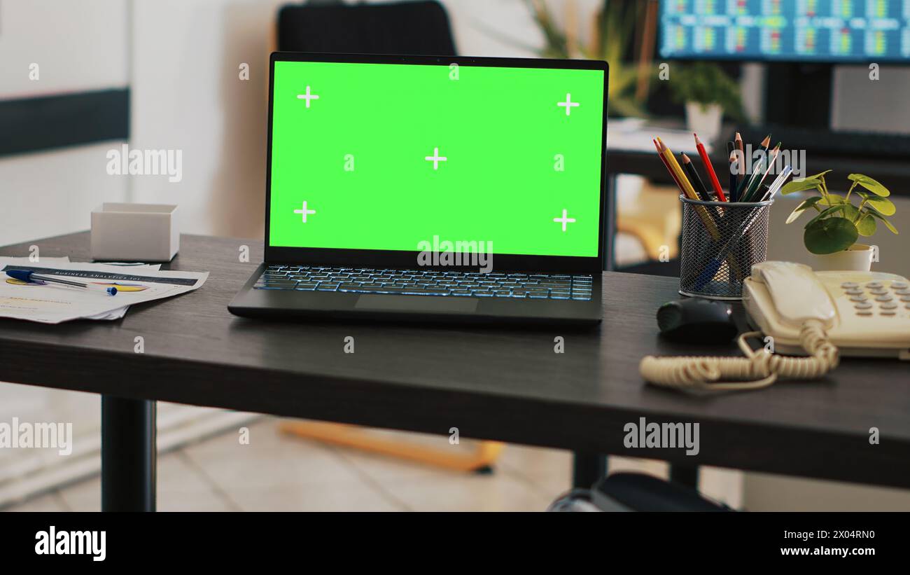Konzentrieren Sie sich auf einen Green-Screen-Laptop auf dem Schreibtisch mit einem Computer im unscharfen Hintergrund, der Börsendaten anzeigt. Nahaufnahme des Modell-Notebooks und der Handelsplattform, die auf dem PC-Bildschirm auf der Rückseite angezeigt werden Stockfoto