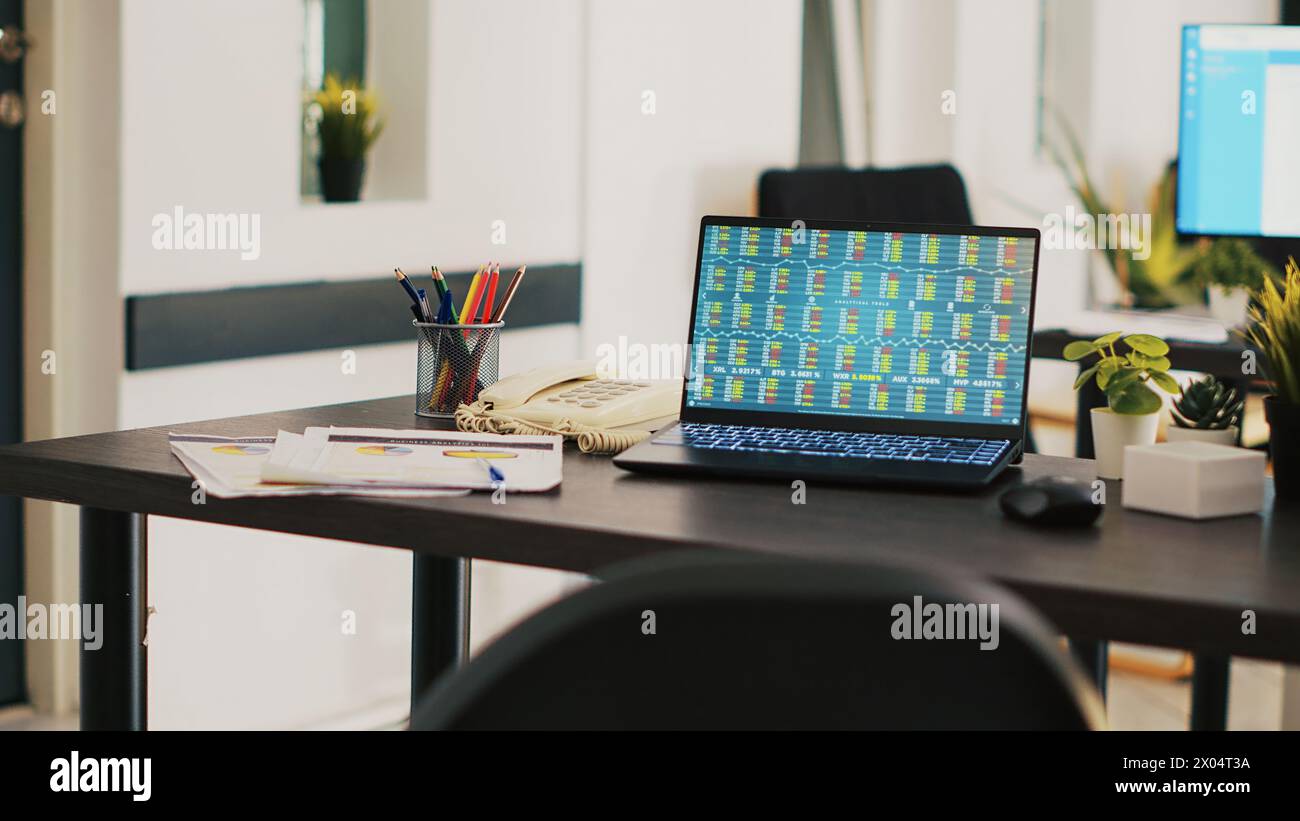 Laptop im Büro des Unternehmens zeigt Börsenwerte in Echtzeit an, die sich ändern. Papierkram auf dem Schreibtisch neben dem Notizbuch mit Forex-Handelsindizes und Tortendiagrammen Stockfoto