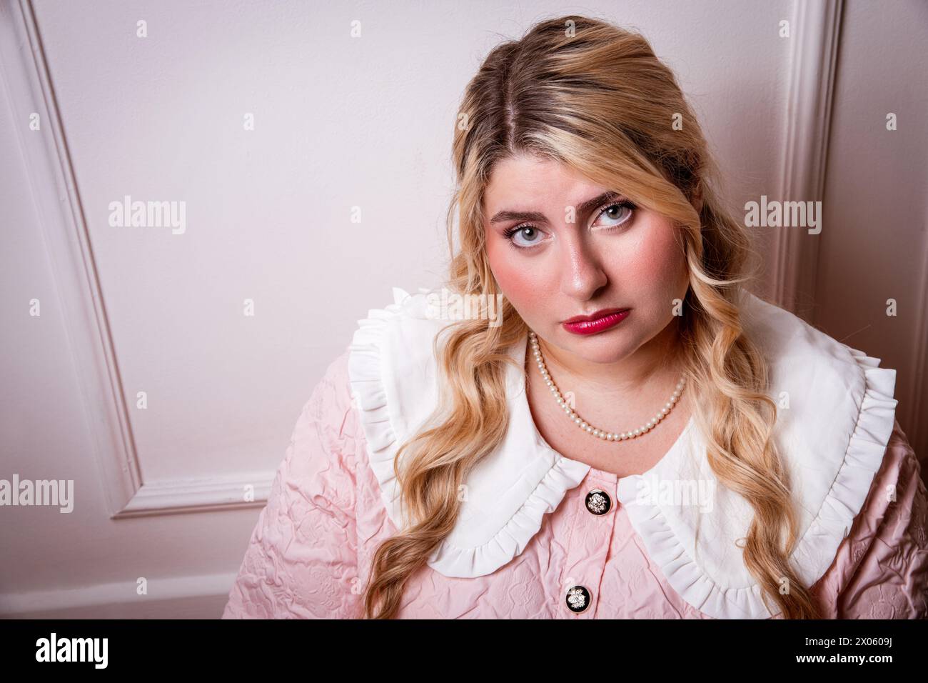 Stilvolle Frau in Übergröße in verärgerter Stimmung, die im Studio eine Perlenkette trägt, die die Kamera vor rosa Hintergrund betrachtet Stockfoto