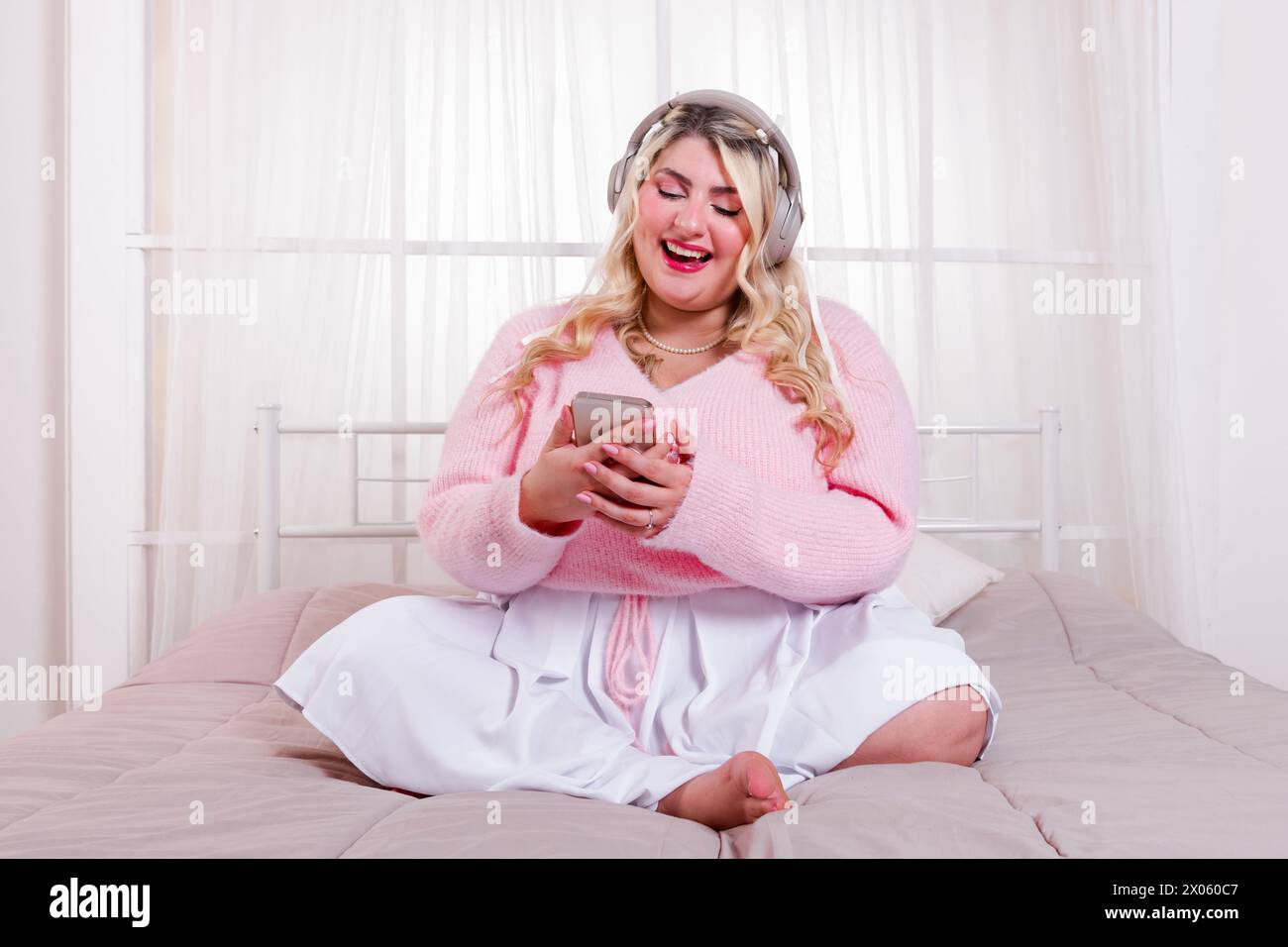 Lächelnde, barfüßige junge Frau in stilvoller Kleidung, die auf dem Bett sitzt, während sie coole Musik mit dem Headset genießt und im hellen Bett auf den Smartphone-Bildschirm blickt Stockfoto