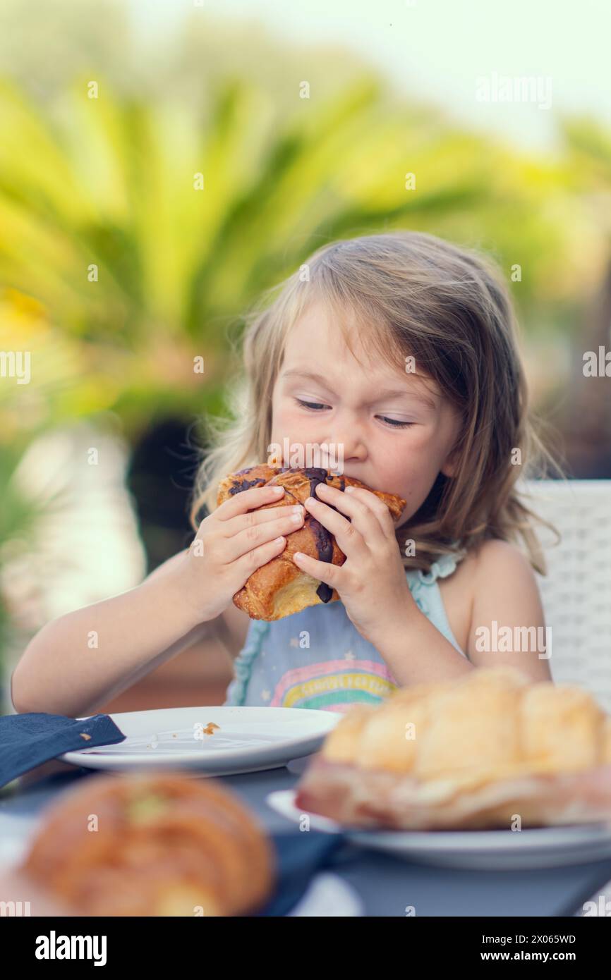 Ein hübsches kleines Mädchen isst ein Croissant mit Schokolade mit Appetit. Das Mädchen frühstückt mit einem Croissant. Das weiße Mädchen isst Gebäck. Nahaufnahme Stockfoto