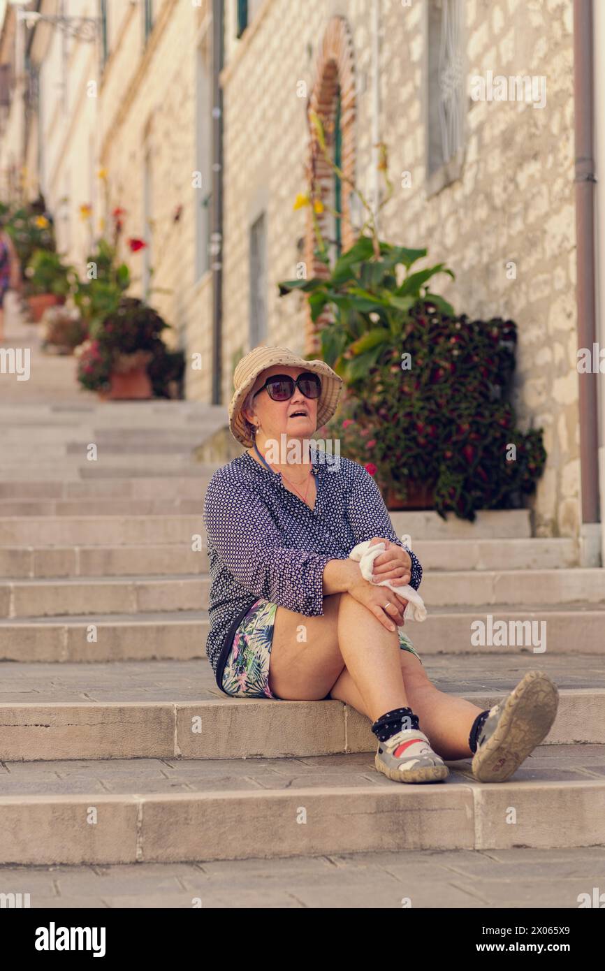 Eine alte Frau mit Hut sitzt auf den Stufen einer langen Treppe. Eine lange Straße mit Stufen ist gesäumt von Topfpflanzen, ein alter Reisender lächelt während er sitzt Stockfoto