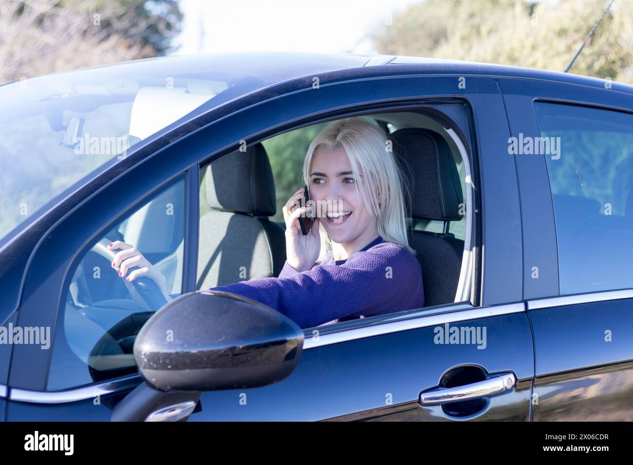 Eine lächelnde blonde Frau in einem lila Pullover, die auf ihrem Handy plaudert, während sie auf dem Fahrersitz eines Autos sitzt Stockfoto