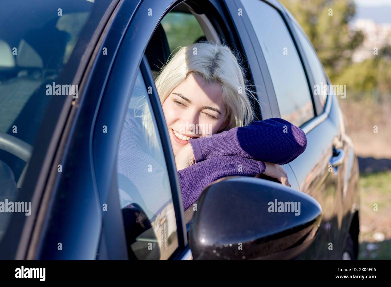 Blonde Frau, die Kopf auf dem Autofenster liegt, die Augen geschlossen, in einem Moment der Entspannung oder Müdigkeit, in einem lila Pullover, in einer ruhigen Atmosphäre Stockfoto