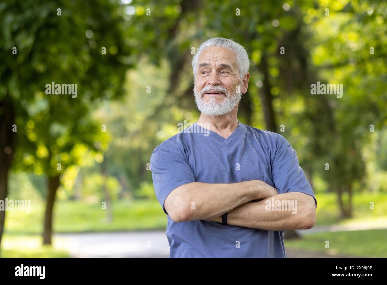 Ein Nahfoto eines älteren grauhaarigen Mannes, der in lässiger Kleidung in der Natur steht, seine Arme auf dem Boden kreuzt und mit einem Lächeln zur Seite blickt. Stockfoto