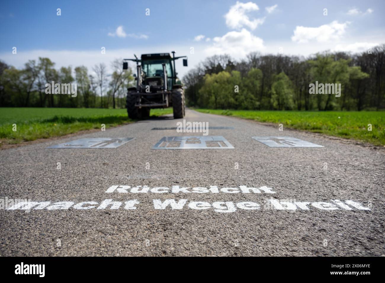 10. April 2024, Bayern, Stadtlauringen: Ein Traktor steht auf einem Weg, auf dem sich mehrere Schablonen für Symbole eines Hundebesitzers, eines Traktorfahrers und eines Radfahrers befinden. Die Schrift „Rücksicht macht Wege breit“ ist bereits in weißer Farbe aufgetragen. Da Konflikte zwischen Landwirten und Freizeitnutzern auf Land- und Forststraßen immer häufiger werden, fordert der Bayerische Bauernverband eine verstärkte Zusammenarbeit mit einer neuen Kampagne und Markierungen auf Feldwegen. Die ersten Logos wurden nun auf einem Weg bei Stadtlauringen im Bezirk SC platziert Stockfoto