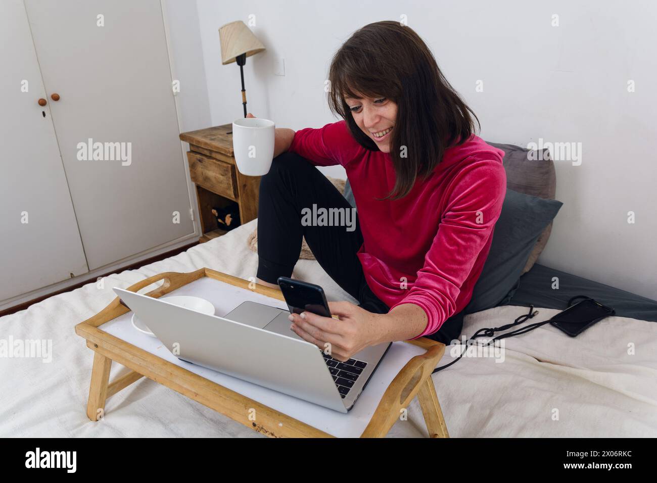 Erwachsene Frau in ihrem Schlafzimmer, die auf dem Bett sitzt, Kaffee glücklich trinkt und mit dem Telefon QR-Code vom Laptop-Bildschirm scannt, um online mit Mobi zu bezahlen Stockfoto