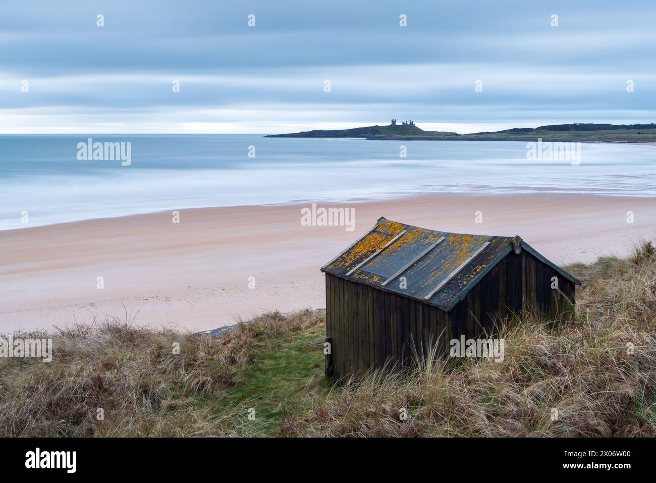 Eine lange Exposition macht die Landschaft, die Embleton Bay an einem grauen Morgen im Frühjahr umgibt, weicher, mit den Ruinen von Dunstanburgh Castle am Horizont. Stockfoto