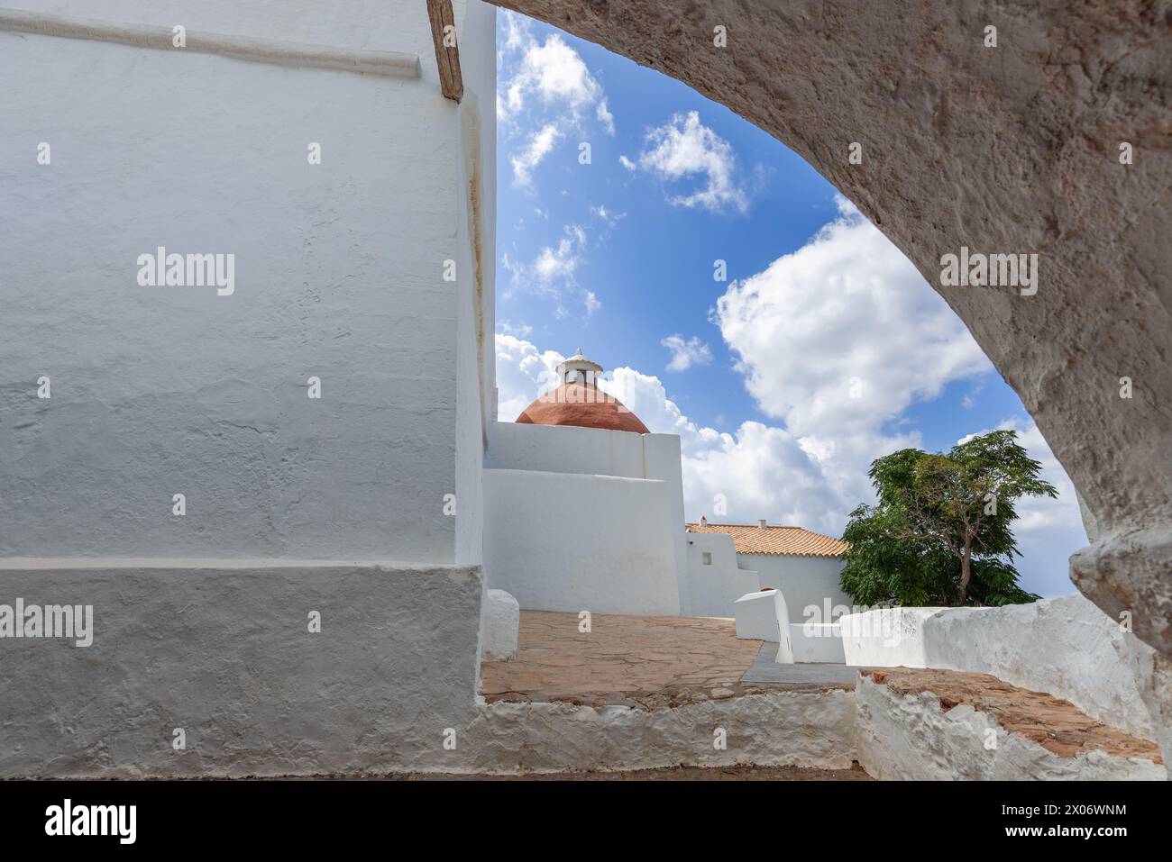 Eingerahmt von einem alten Steinbogen erhebt sich die Terrakotta-Kuppel der Kirche Puig de Missa in Santa Eulalia, Ibiza, über dem blauen Himmel. Weiße Wände und eine üppige tr Stockfoto