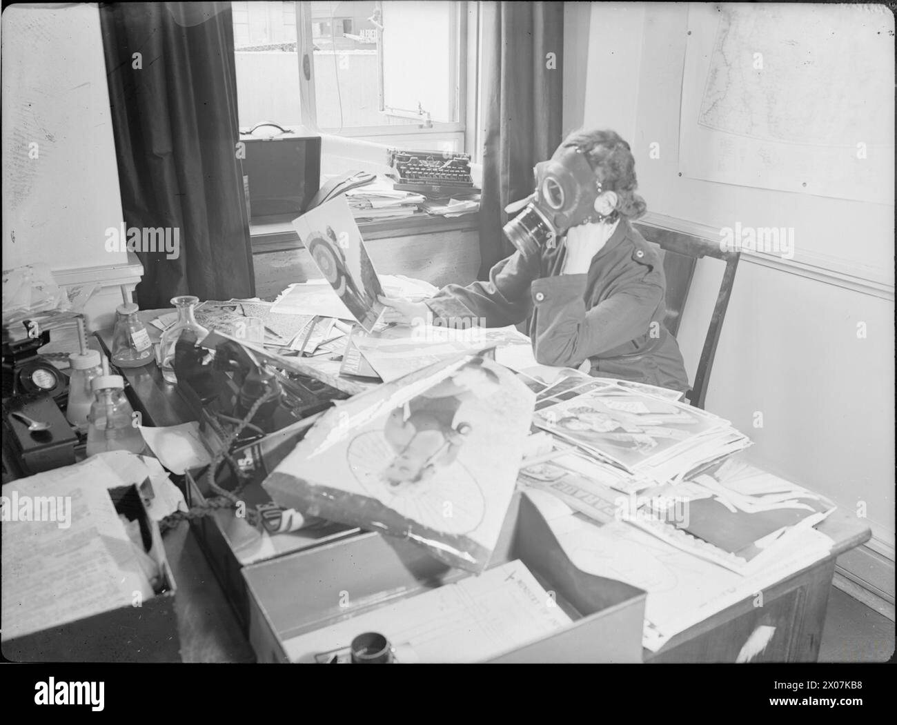 LUFTANGRIFFE IN GROSSBRITANNIEN: MITARBEITER DES INFORMATIONSMINISTERIUMS TRAGEN GASMASKEN ZUR ARBEIT, LONDON, ENGLAND, 1941 - Miss Marjorie Lloyd von der Nahost-Abteilung des Informationsministeriums sitzt mit einer zivilen Gasmaske an ihrem Schreibtisch. Ihre Aufgabe ist es, Fotos in London für den Versand an das Magazin „Parade“ auszuwählen Stockfoto