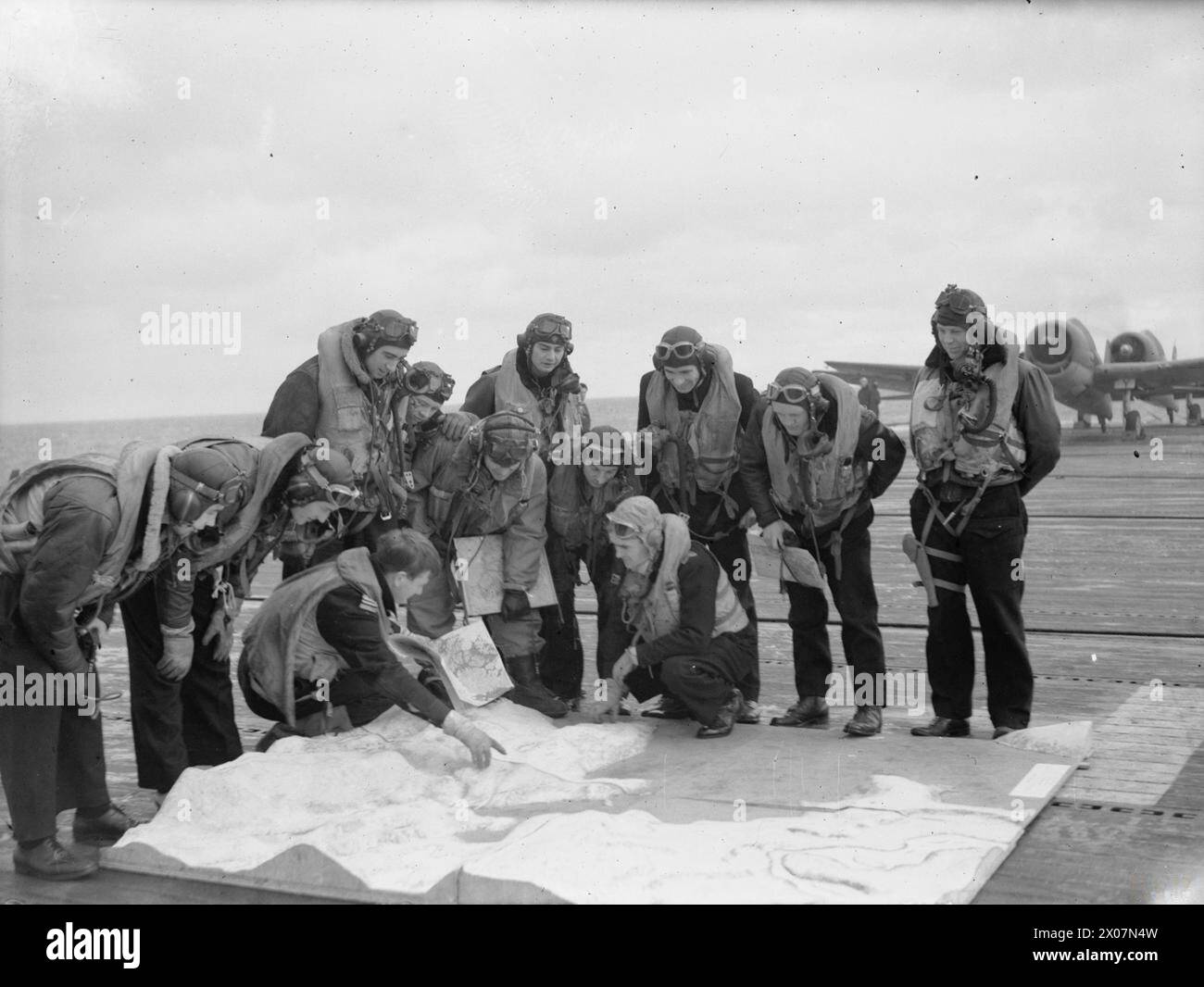 Grumman Hellcat-Piloten an Bord der HMS Emperor studieren vor ihrem Angriff ein Modell des deutschen Schlachtschiffs Tirpitz im Alten Fjord, wobei zwei Flugzeuge in der Ferne sichtbar sind. Stockfoto