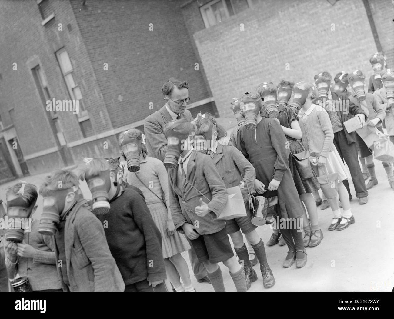 LONDON SCHOOLS IN WARTIME: SCHULLEBEN IN LONDON, ENGLAND, 1941 – Schulkinder stehen auf dem Spielplatz während einer Gasmasken-Übung an der Old Woolwich Road School, Greenwich. Der Lehrer passt den Gurt an einer der Kindermasken an Stockfoto