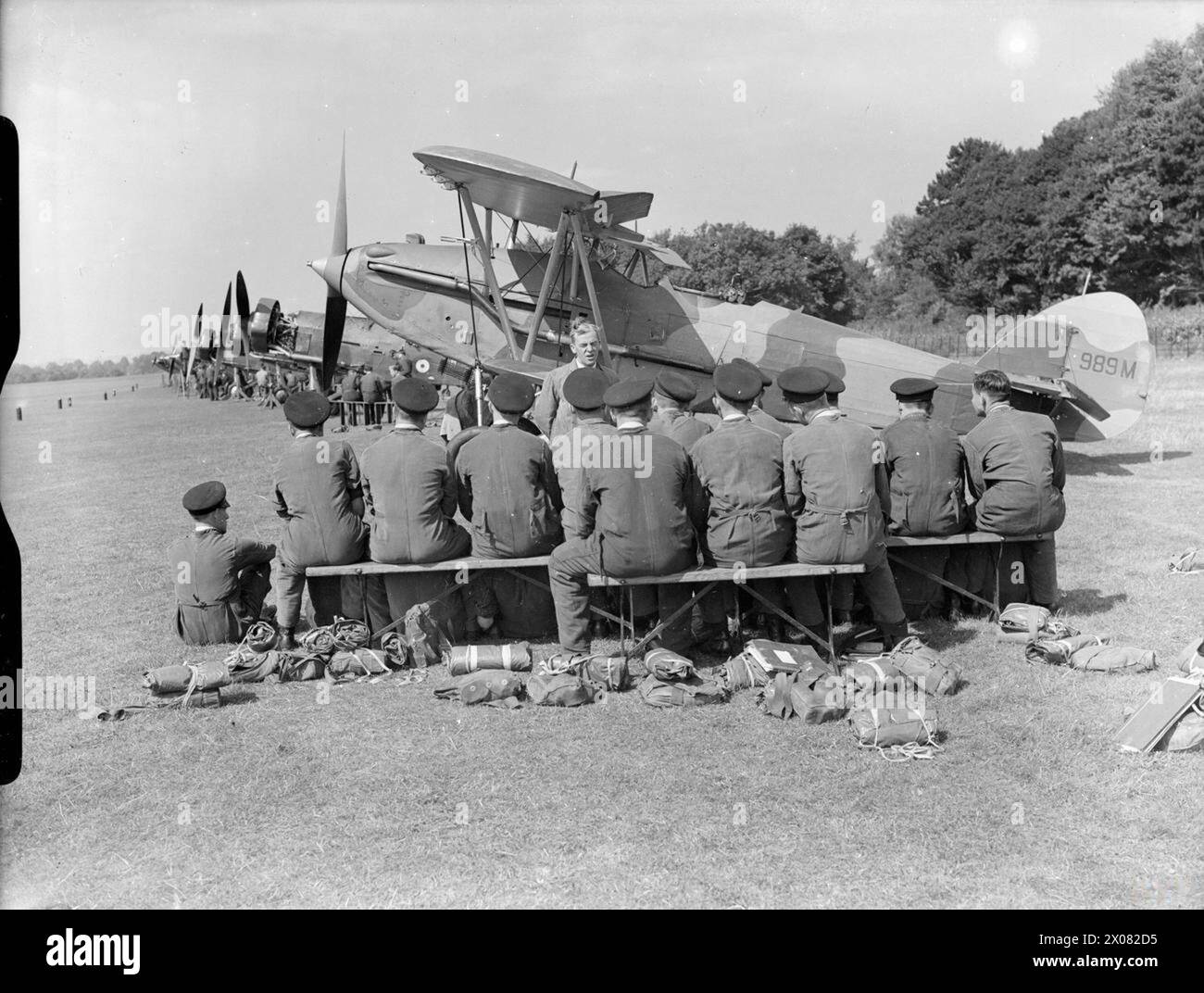 DAS TECHNISCHE AUSBILDUNGSKOMMANDO DER ROYAL AIR FORCE, 1940-1945. - Flugzeuglehrlinge der Schule für technische Ausbildung Nr. 1 hören einen Vortrag über die Wartung von Flugzeugen im Feld, vor einer Reihe von Lehrflugzeugen auf dem Flugplatz in Halton, Buckinghamshire. 989M ist eine Hawker Audax Mark I, ehemals K3057 der No. 2 Squadron RAF, die 1937 in den Flugzeugstatus der Royal Air Force, School, Technical Training, 5, Locking reduziert wurde Stockfoto