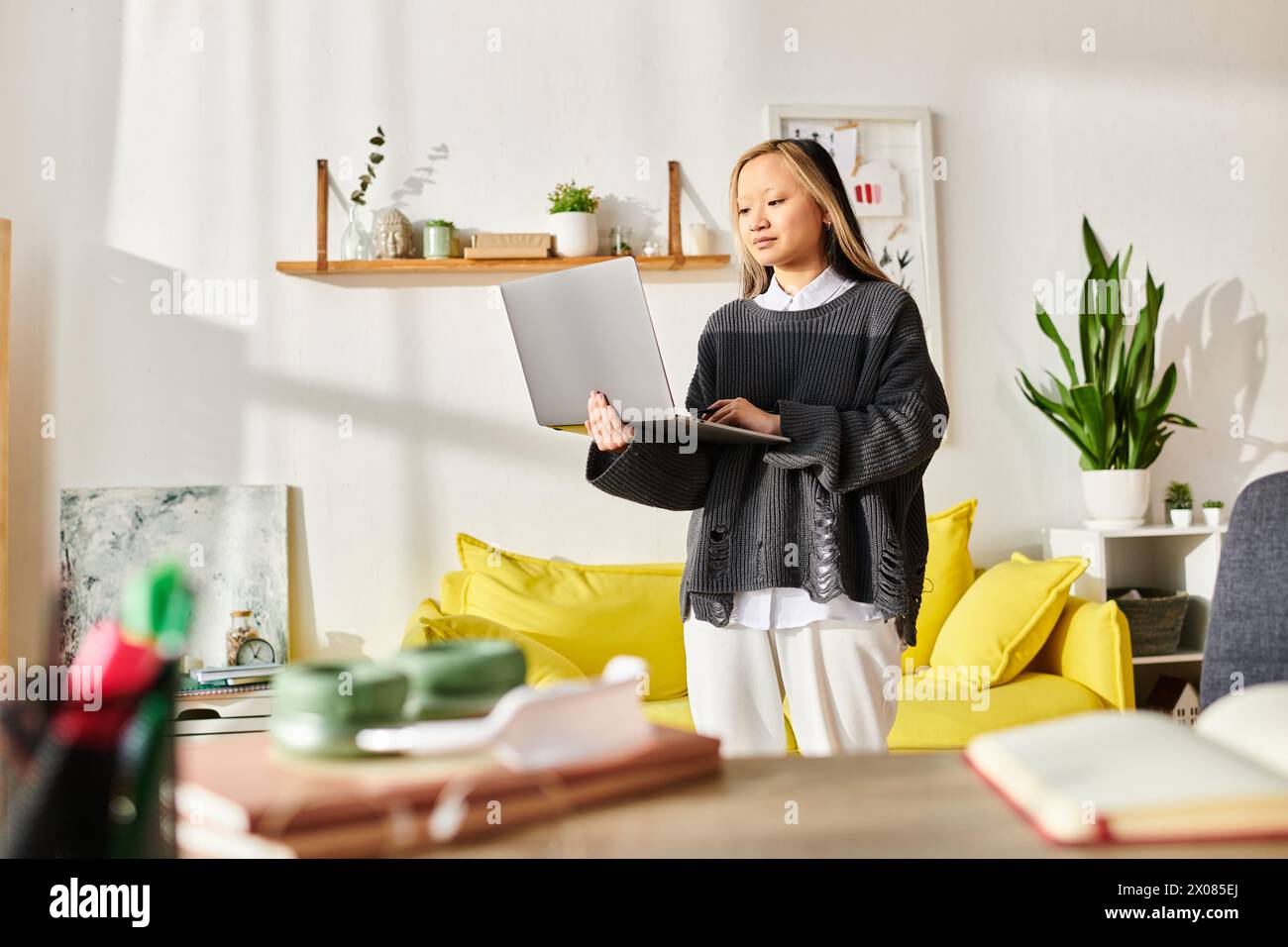 Eine junge asiatische Frau steht elegant in einem modernen Wohnzimmer, vertieft in E-Learning, während sie einen Laptop in der Hand hält. Stockfoto