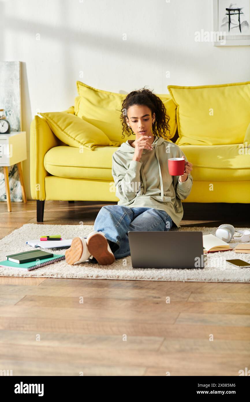 Eine junge Frau sitzt auf dem Boden und hält sich anmutig an eine warme Tasse Kaffee, während sie zu Hause auf ihrem Laptop studiert. Stockfoto