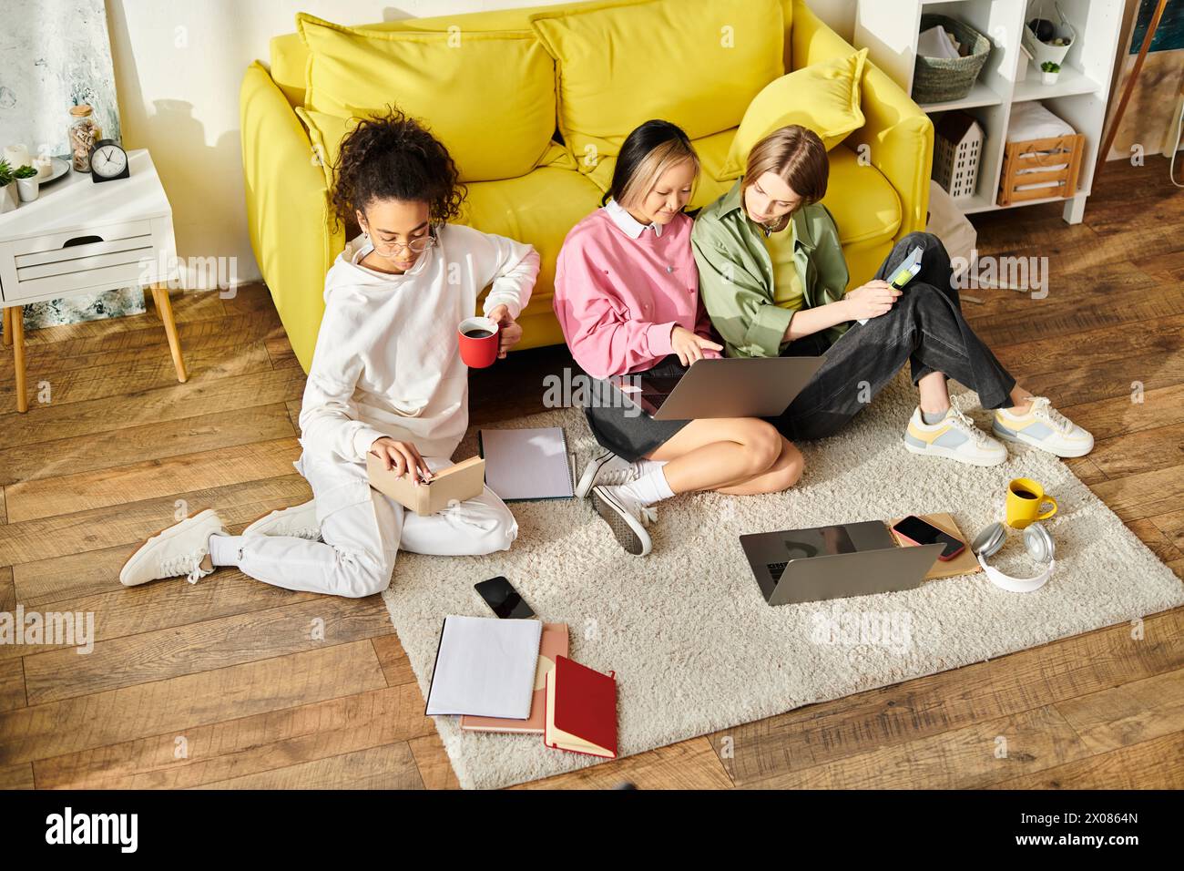Drei verschiedene Teenager-Mädchen sitzen auf dem Boden vor einer gelben Couch, vertieft in das gemeinsame Lernen. Stockfoto