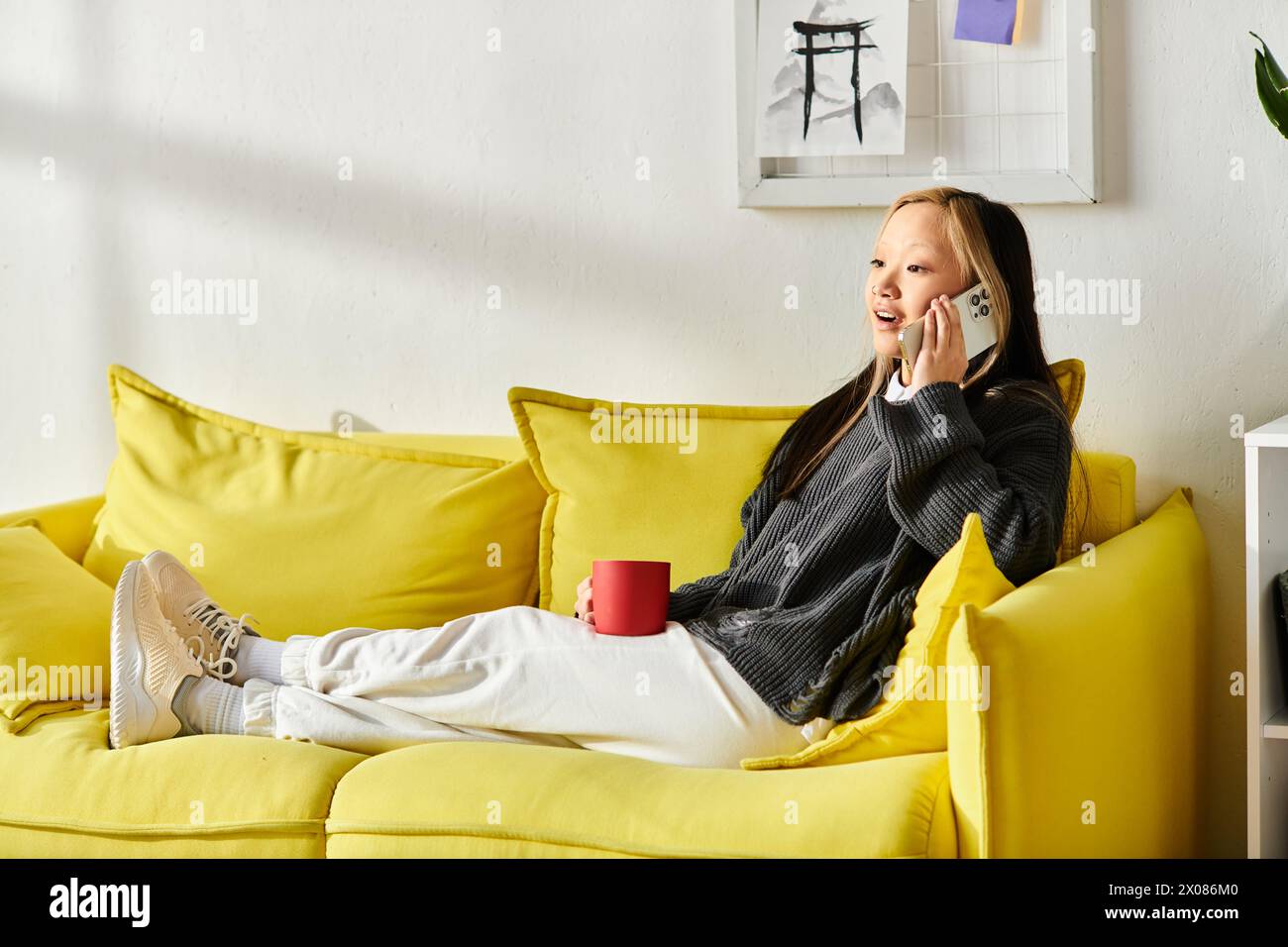 Eine junge asiatische Frau sitzt bequem auf einer gelben Couch und führt auf ihrem Handy ein lebhaftes Gespräch. Stockfoto
