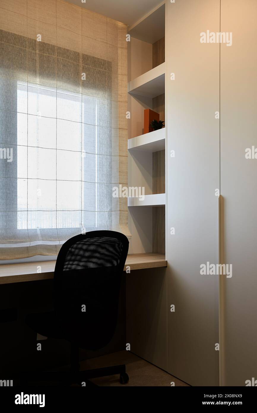Moderne Einrichtung für das Home Office mit einem eleganten Schreibtisch, ergonomischem Stuhl und eingebauten Regalen unter natürlichem Licht durch ein Fenster mit transparenten Vorhängen Stockfoto