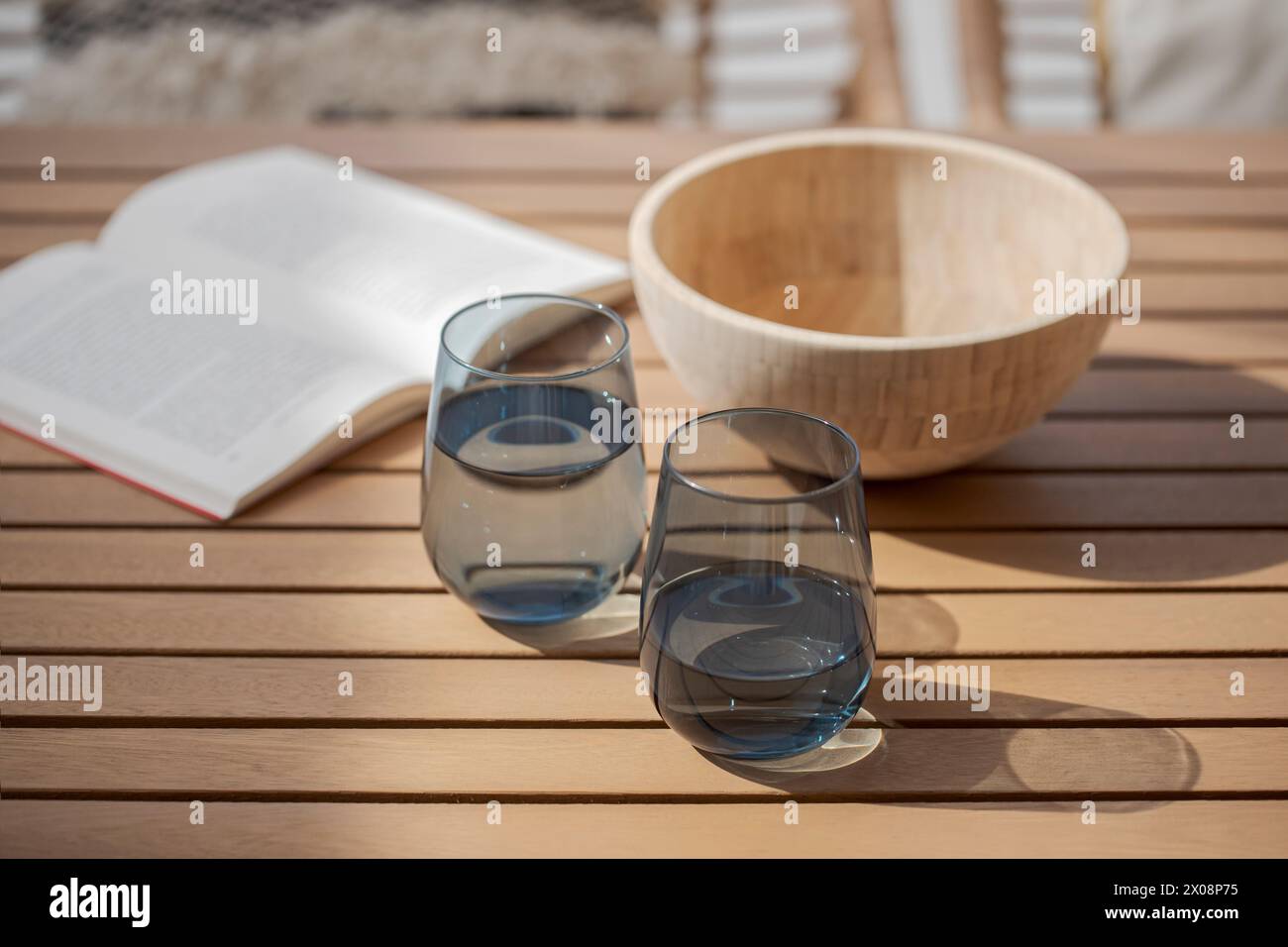 Eine gemütliche Sommerszene mit einem offenen Buch und zwei stiellosen Weingläsern gefüllt mit Wasser auf einem hölzernen Terrassentisch, begleitet von einem leeren Holzbogen Stockfoto