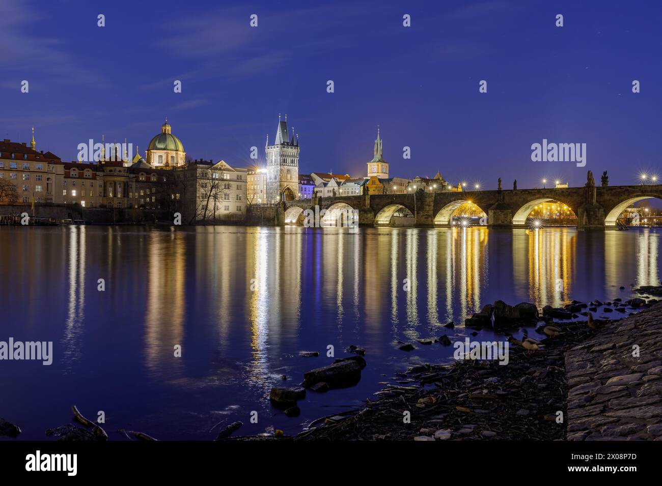 Eine ruhige Dämmerungsszene, in der die beleuchtete Karlsbrücke auf der Moldau in Prag reflektiert wird, mit historischen Gebäuden im Hintergrund Stockfoto