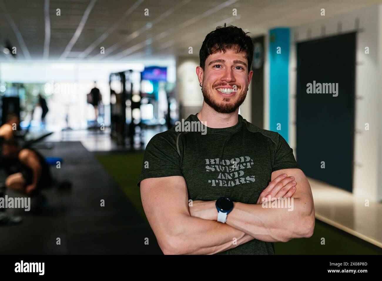 Ein fröhlicher junger Mann steht mit überkreuzten Armen in einem Fitnessstudio, trägt sportliche Kleidung und eine Smartwatch und strahlt Selbstvertrauen und Fitness-Vibes aus. Stockfoto