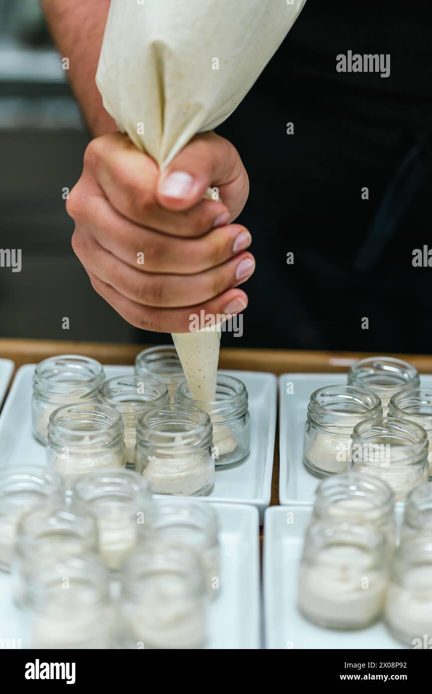 Nahaufnahme der Hände des anonymen Küchenchefs, während sie geschickt Vanillecreme in kleine Gläser spritzen und ein Gourmet-Dessert zubereiten Stockfoto
