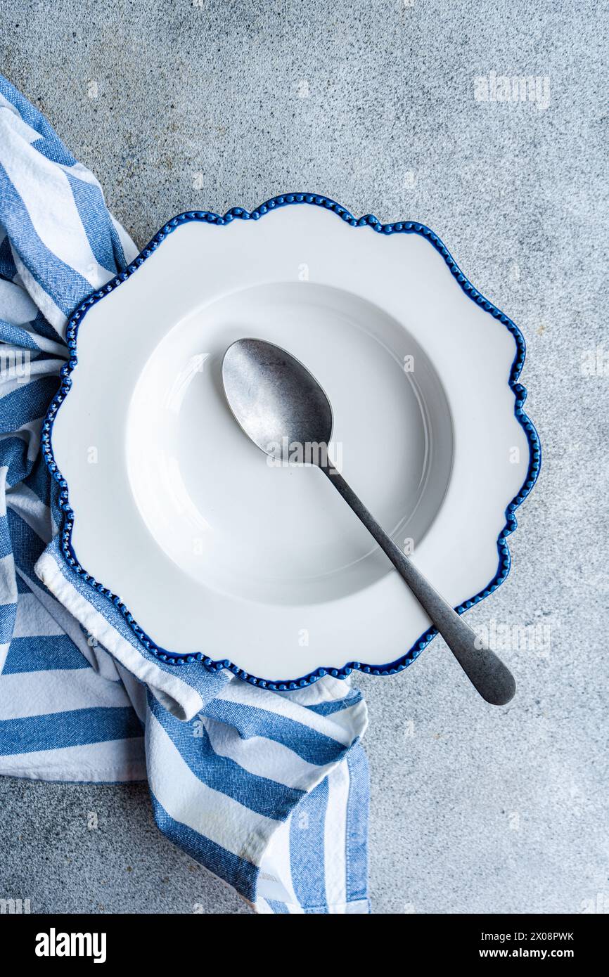 Ein eleganter weißer Teller mit gewellten blauen Kanten, begleitet von einem Vintage-Löffel, auf einem Betonhintergrund, gewickelt in ein gestreiftes blaues Geschirrtuch Stockfoto