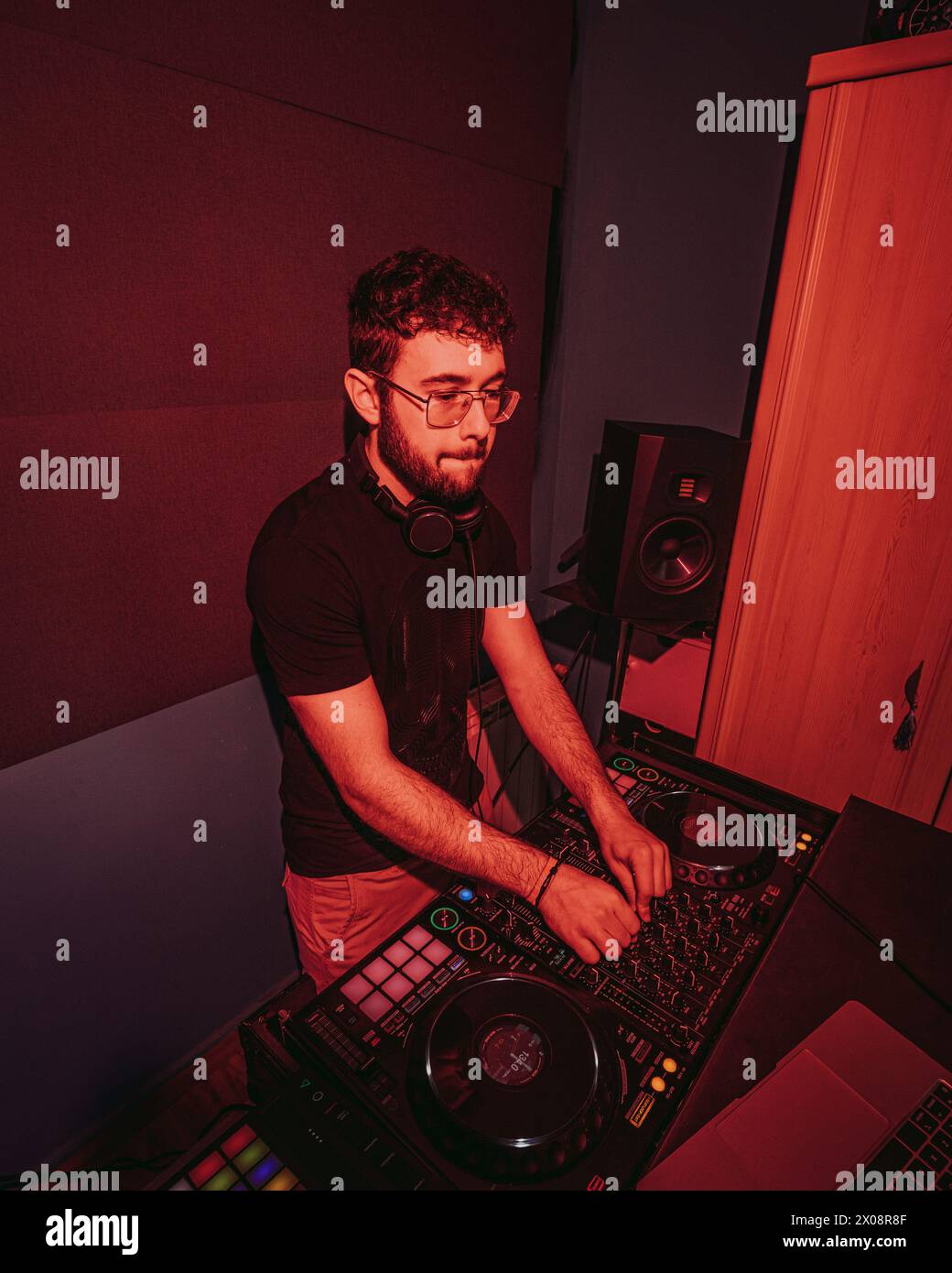 Ein fokussierter DJ, der Kopfhörer trägt, mischt Songs auf einem professionellen DJ-Controller, beleuchtet durch rote Umgebungsbeleuchtung in einem Club-ähnlichen Studio-Setting Stockfoto