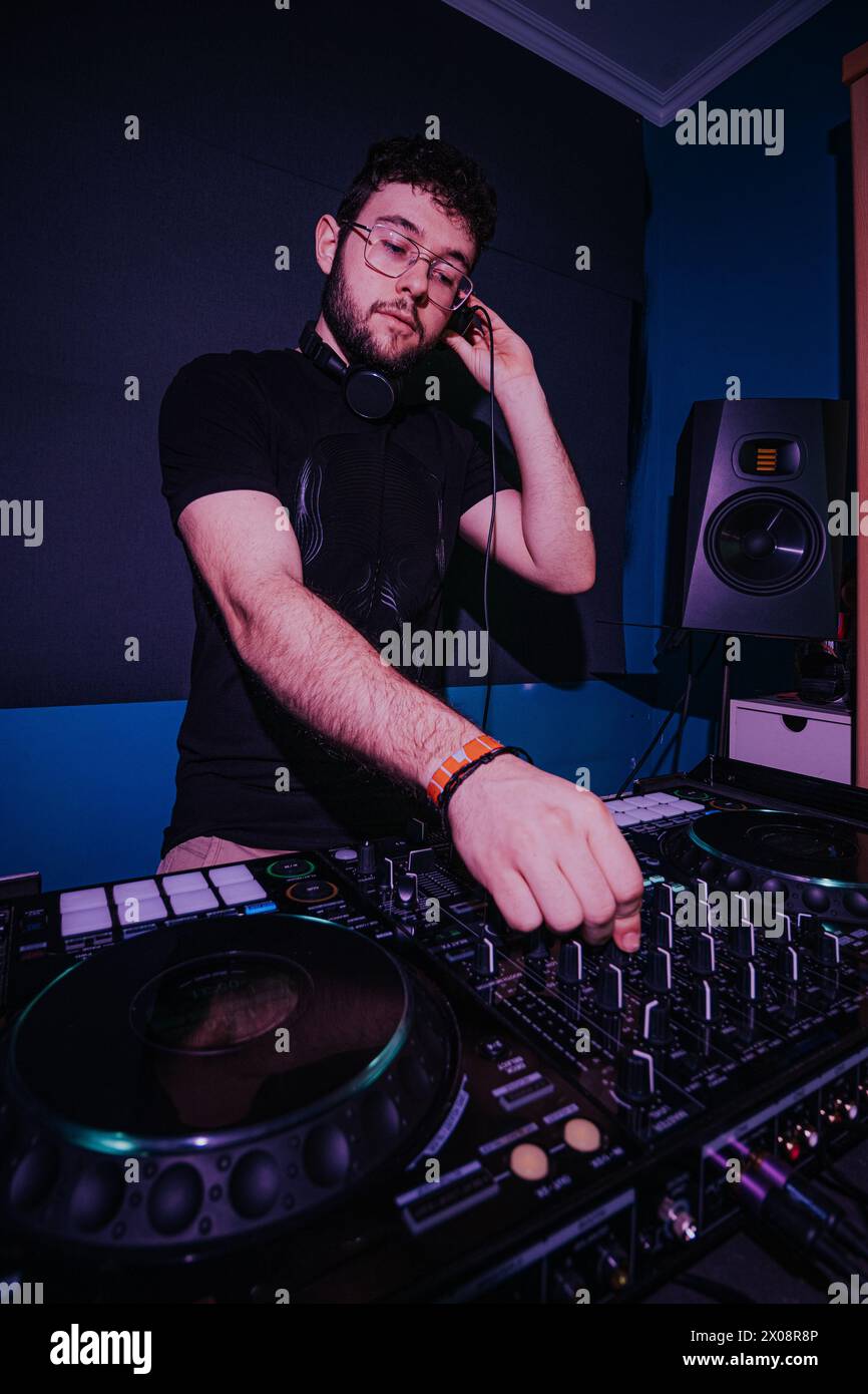 Ein fokussierter männlicher DJ passt seine Kopfhörer an und mischt Musik auf einer professionellen DJ-Konsole in einem schwach beleuchteten Raum mit stimmungsvoller Beleuchtung Stockfoto