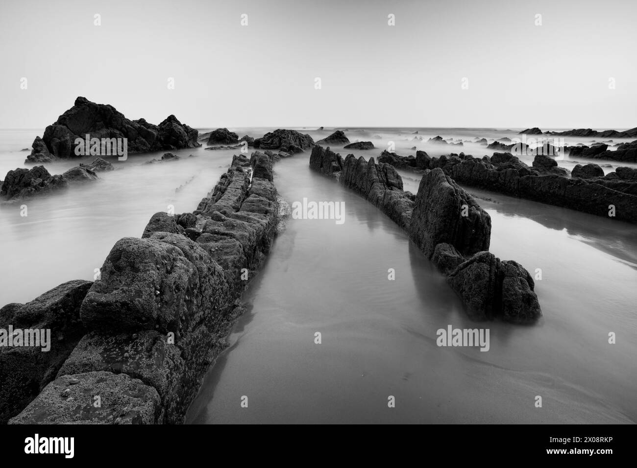 Die strukturierten Felsen von Playa de Barrika stehen in dieser dramatischen schwarz-weißen Küstenszene in der Nähe von Bilbao stark gegen das glatte Meer Stockfoto