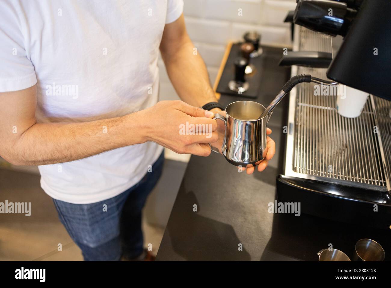 Ein Barista in lässiger Kleidung schäumt Milch mit einer professionellen Espressomaschine auf und konzentriert sich dabei auf den glänzenden Metallkrug. Stockfoto