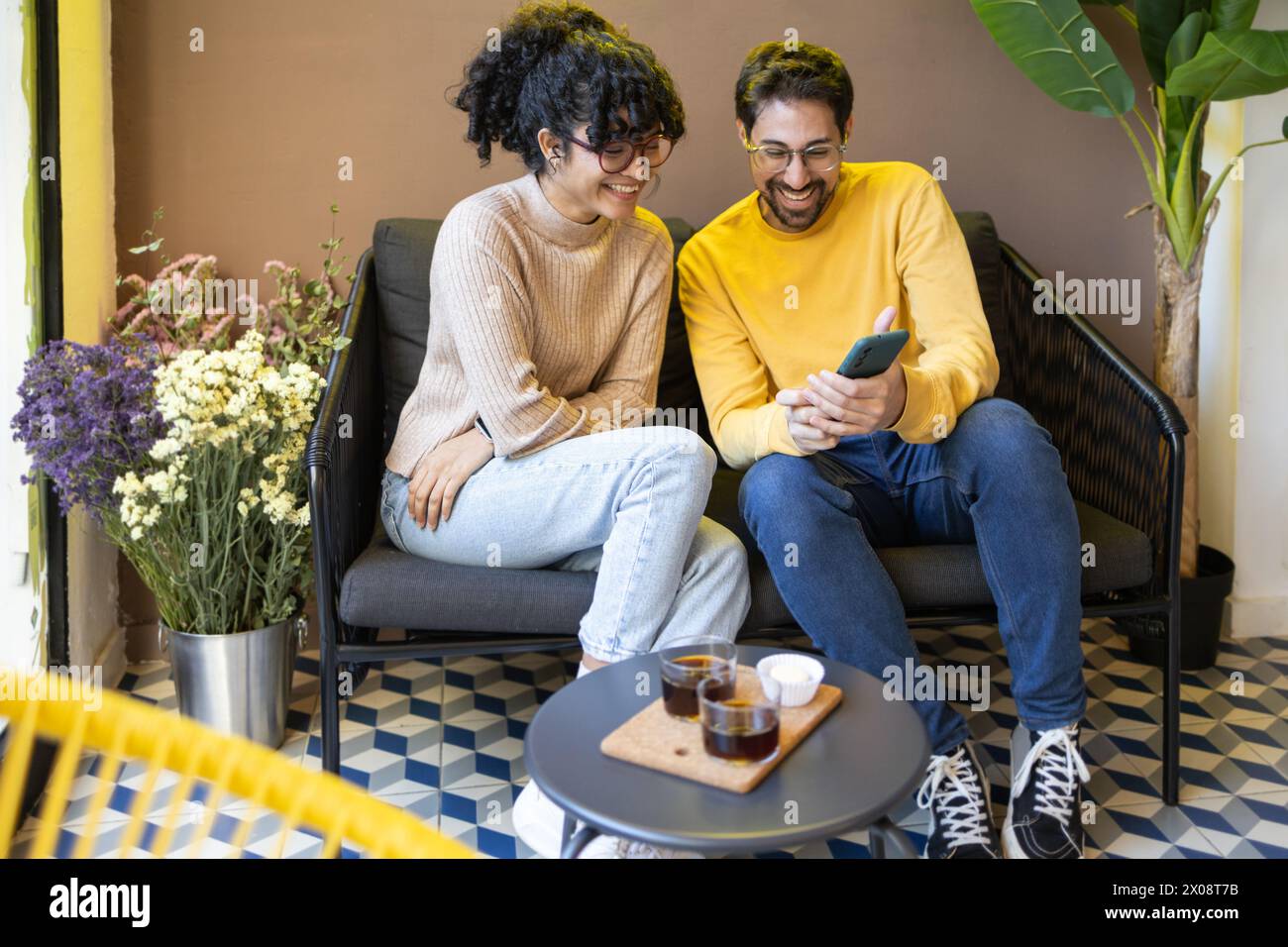 Ein fröhliches, multiethnisches Paar sitzt eng auf einer Couch, vertieft in einem Smartphone-Bildschirm, lacht in einer gemütlichen Innenumgebung mit dekorativen Blumen nea Stockfoto