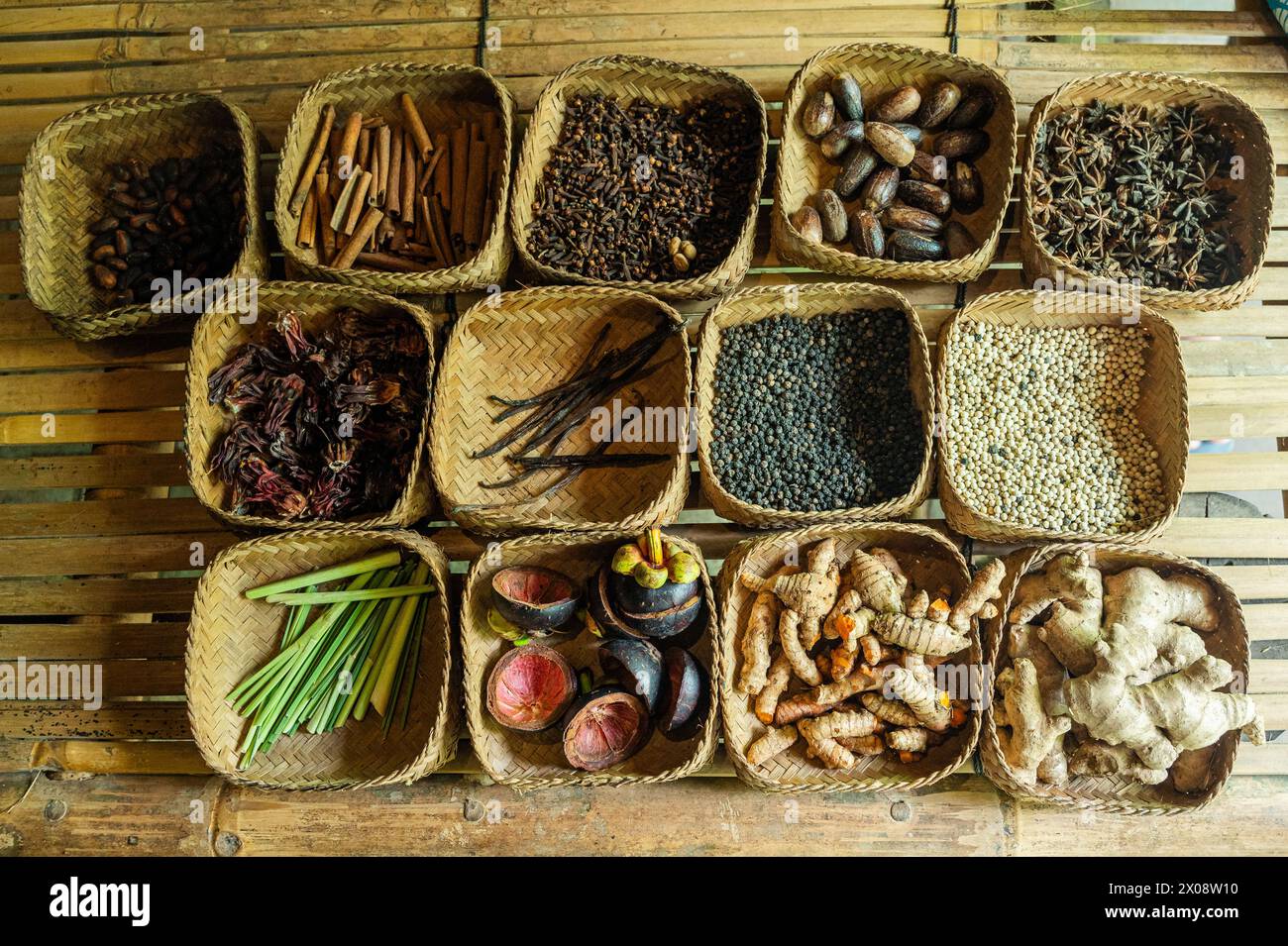 Verschiedene Gewürze in gewebten Körben, traditionell auf Bali, mit Zimt, Muskatnuss, schwarzem Pfeffer, Vanille und mehr auf einem rustikalen Holztisch. Stockfoto